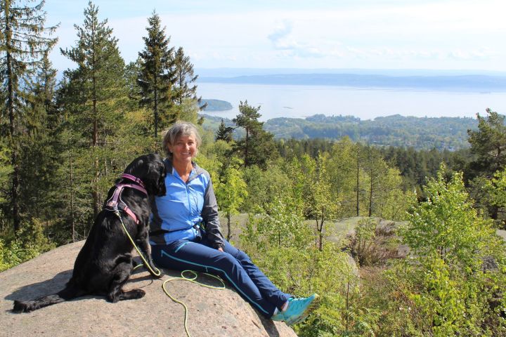 En dame og en hund på en fjelltopp med utsikt mot en fjord i bakgrunnen. Pent vær.