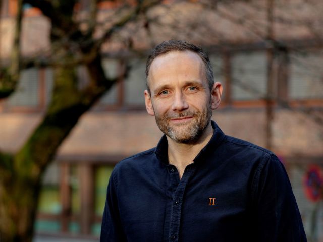 Portrett av pen, smilende mann med skjegg og blå skjorte