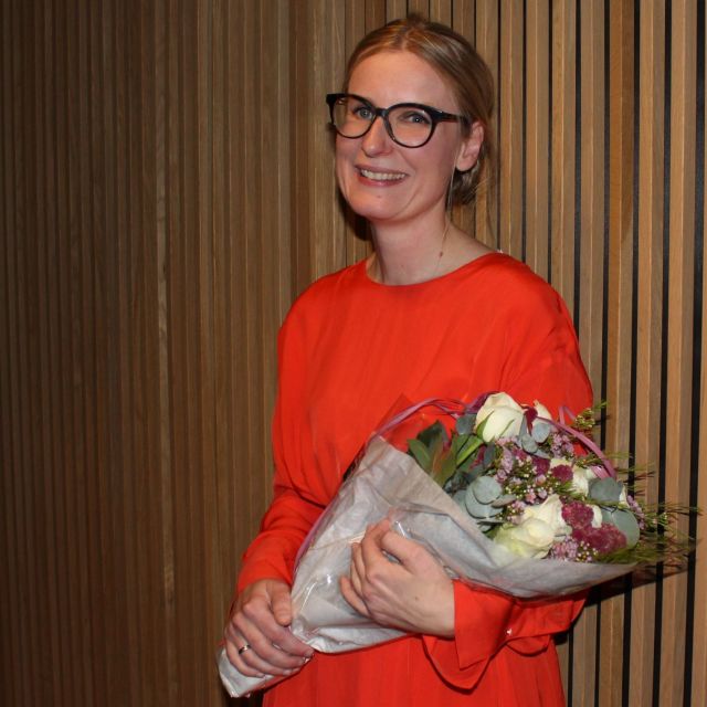 Smildene, lys dame i orange kjole og med blomsterbukett på armen.