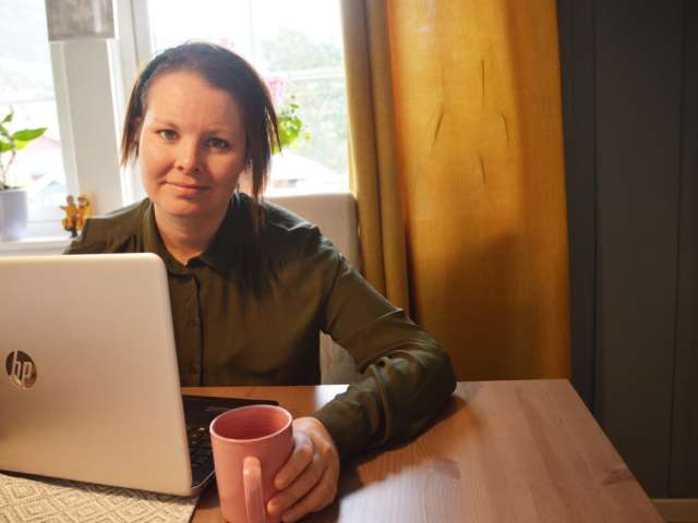 Dame som sitter foran en bærbar PC med kaffekopp i hånden.